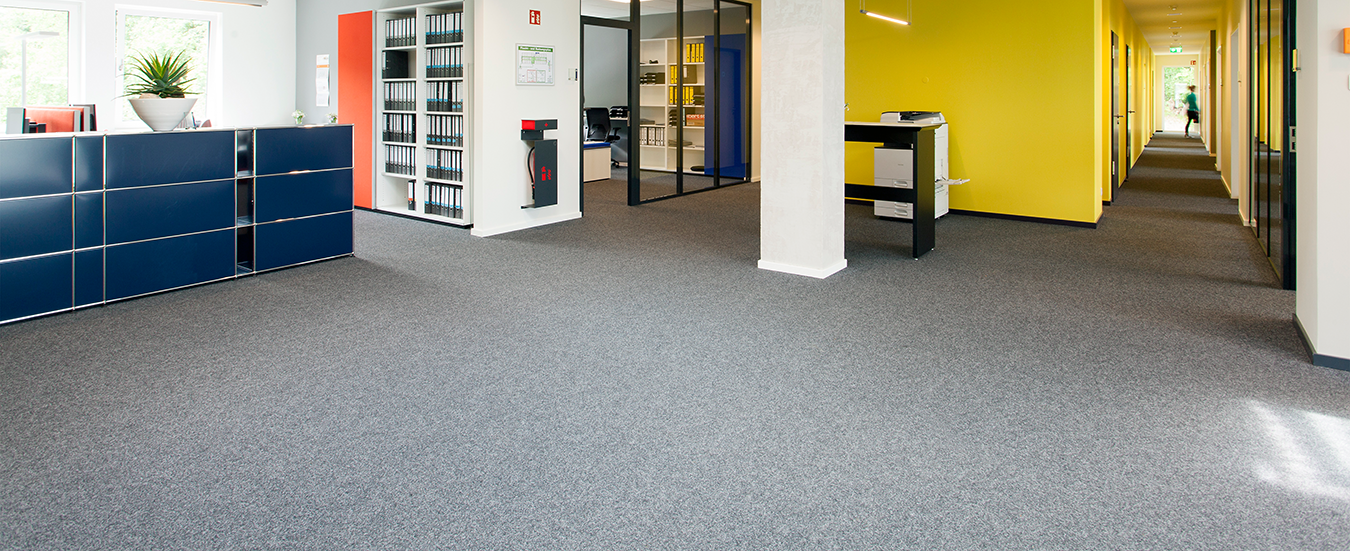 Boenbeläge Hespenheide 1 Moderner Bürobereich mit grauem Teppichboden, Empfangstresen und farblich abgesetztem Flur.