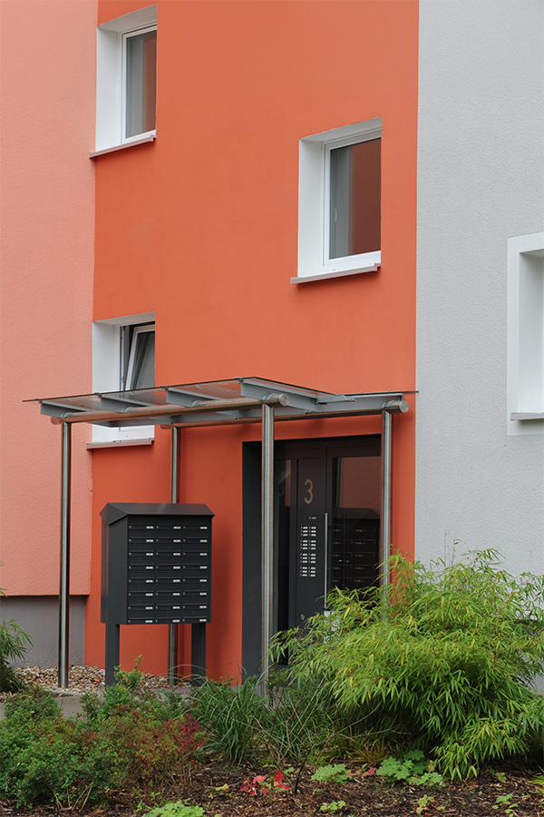 Moderner Eingangsbereich mit farbig verputzter Fassade, Glasvordach und Briefkastenanlage.