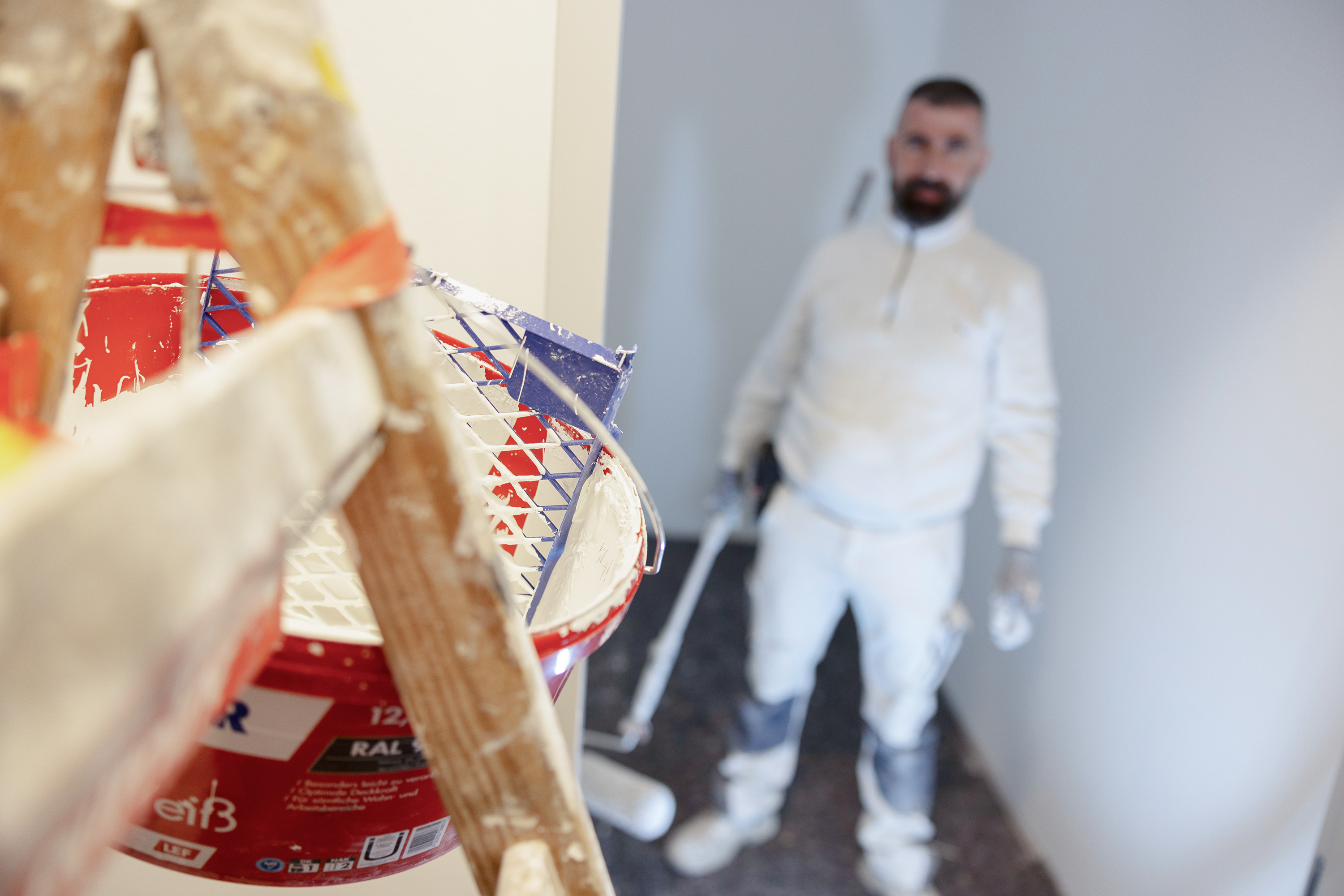 Maler arbeitet in Innenraum mit Farbrolle und Farbkübel im Vordergrund während Renovierungsarbeiten.