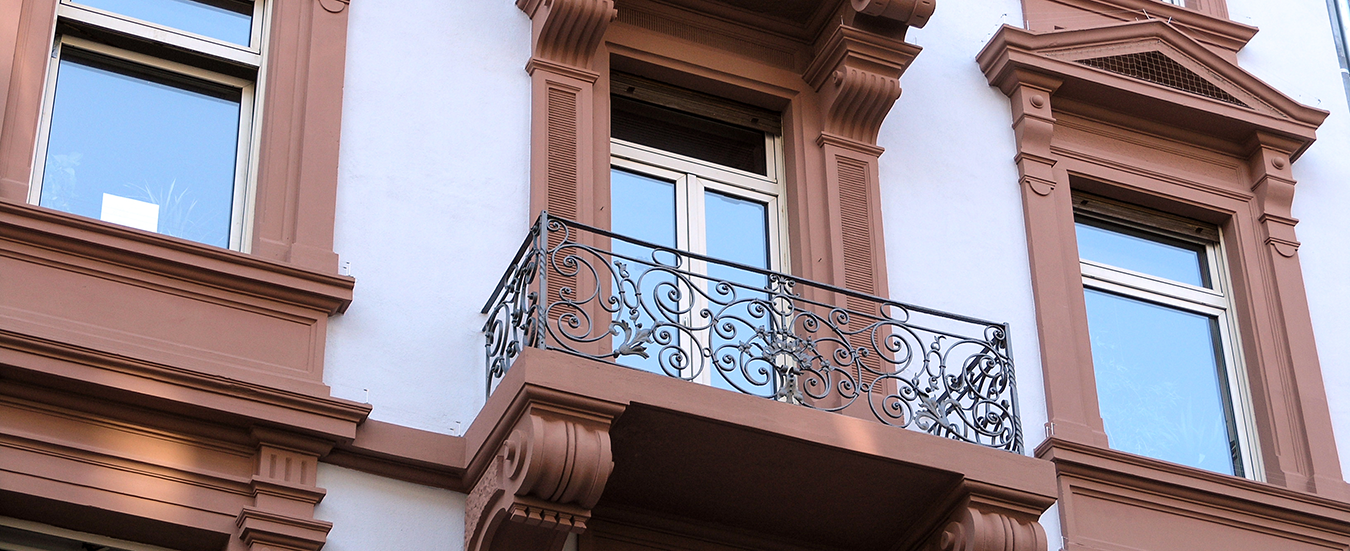 WichsleFassadengestaltung Teaser Detail einer aufwendig gestalteten Altbaufassade mit Balkon, dekorativen Stuckelementen und aufwendig gestrichenen Fensterrahmen.