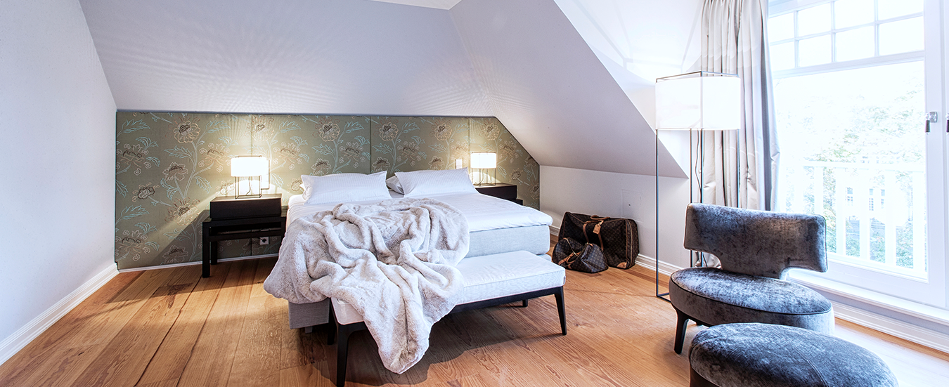 Wichels Malerarbeiten Teaser Modernes Schlafzimmer mit Doppelbett, gepolstertem Hocker, Holzfußboden, Fensterfront und dekorativer Tapetenwand.