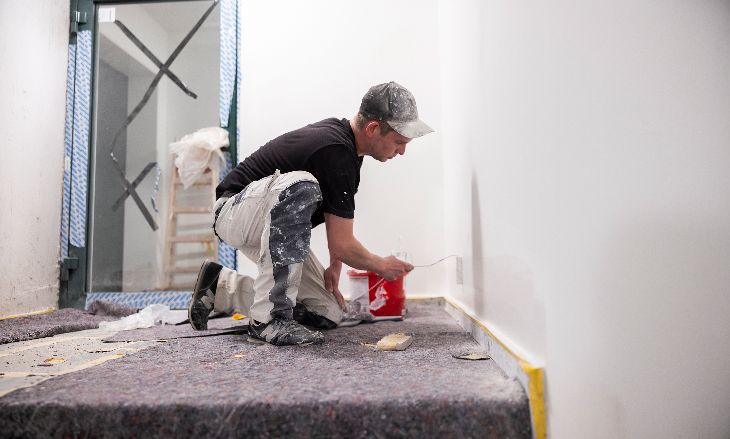 Wichels Maler Leistungen Handwerker streicht Innenwand mit Farbrolle beim Renovieren, mit Farbeimer und Schutzabdeckung auf dem Boden.